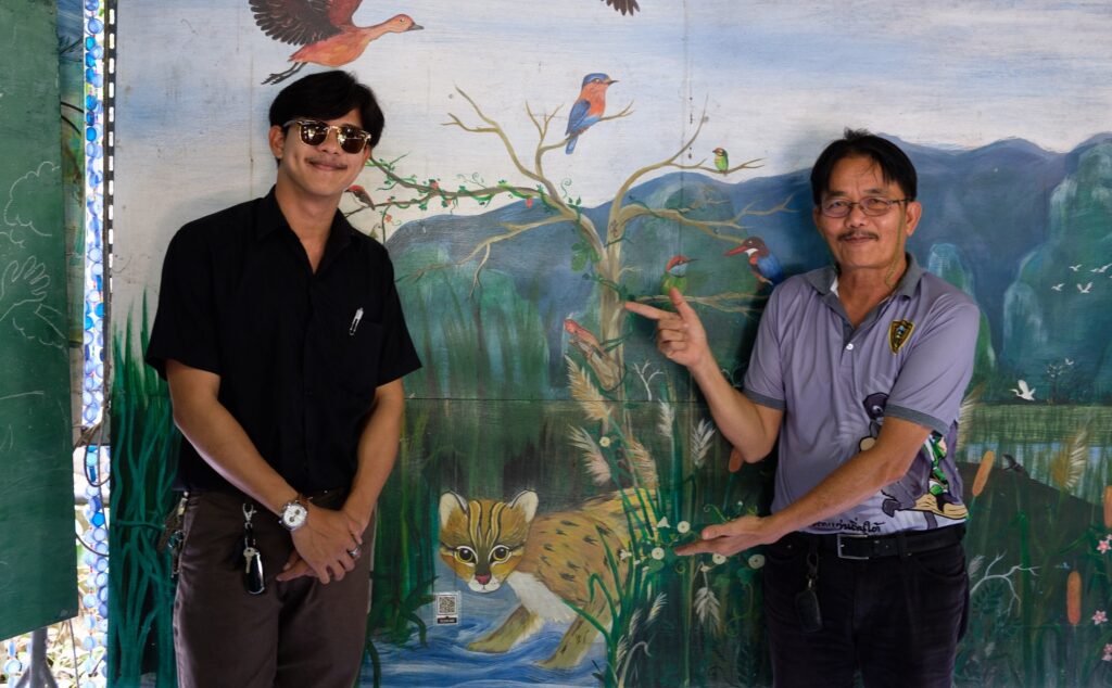 Supoj Sukpat (right), a school teacher and leader of the Rak Thung youth conservation group shows a mural of a fishing cat painted by Pisan Khamhom (left), a former conservation group student and now a teacher at the Sam Roi Yot school.