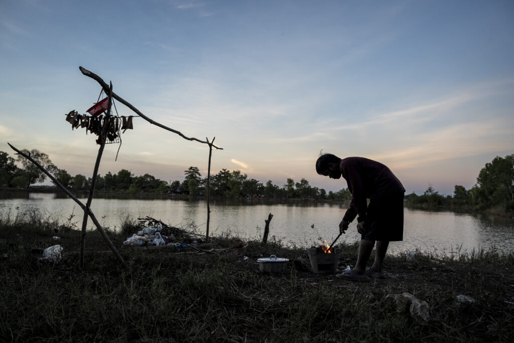 A man stokes a cooking fire in front of the pond at Chroy Neang Ngourn pagoda, where thousands of displaced families temporarily reside, in Cambodia's Siem Reap province on December 15, 2025. (Roun Ry/Mekong Independent)