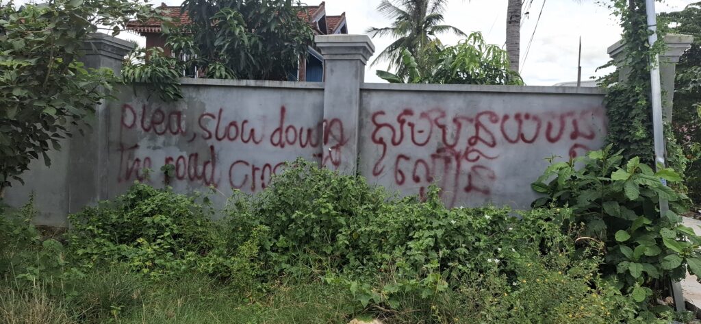 A spray-painted message on a house's fence, asking drivers to slow down in English and Khmer, on the Takeo province road leading to Mango 2 Park, in Cambodia on August 13, 2025. (Danielle Keeton-Olsen/Mekong Independent)