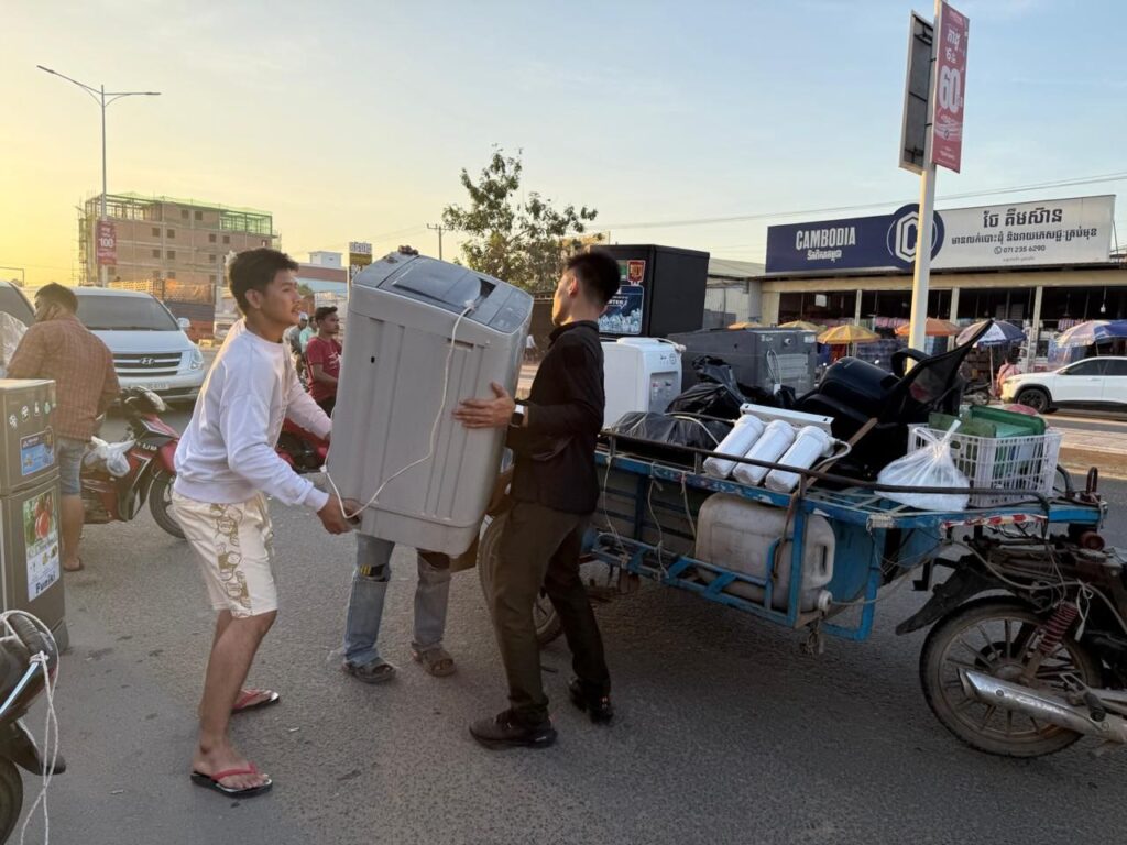 Three men load a washing machine into a packed motorbike trailer parked along National Road 1 in Cambodia's Bavet city on February 1, 2026. (Mech Dara/Mekong Independent)