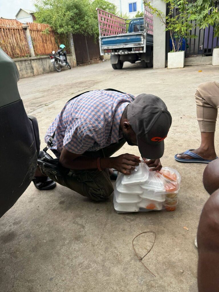 A Ugandan man opens a bag filled with packaged meals bought by the Caritas shelter for the foreign nationals who sit waiting outside the NGO's Phnom Penh office on February 27, 2026. (Mech Dara/Mekong Independent/Creative Commons)