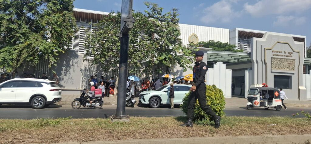 A military police offer walks on the boulevard in front of the National Bank of Cambodia headquarters as clients of Huione Pay and H Pay wait to raise their complaints on April 7, 2026. (Danielle Keeton-Olsen/Mekong Independent/Creative Commons)